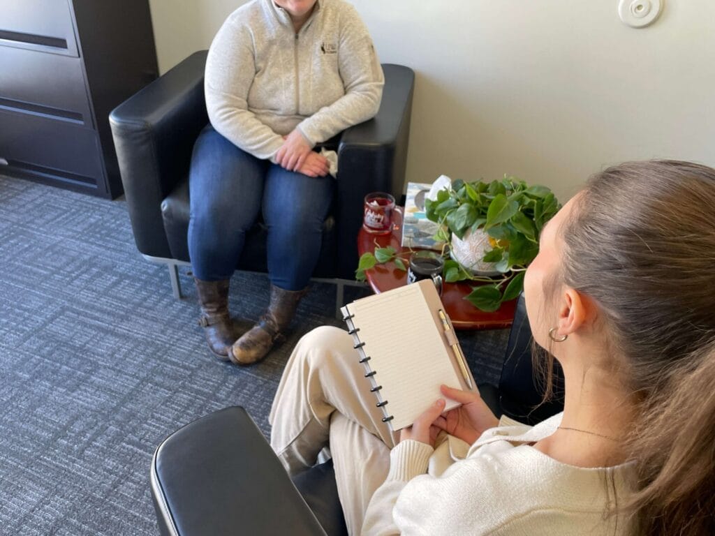 A therapist holding a notepad while listening to a patient seated across from her in an office setting.