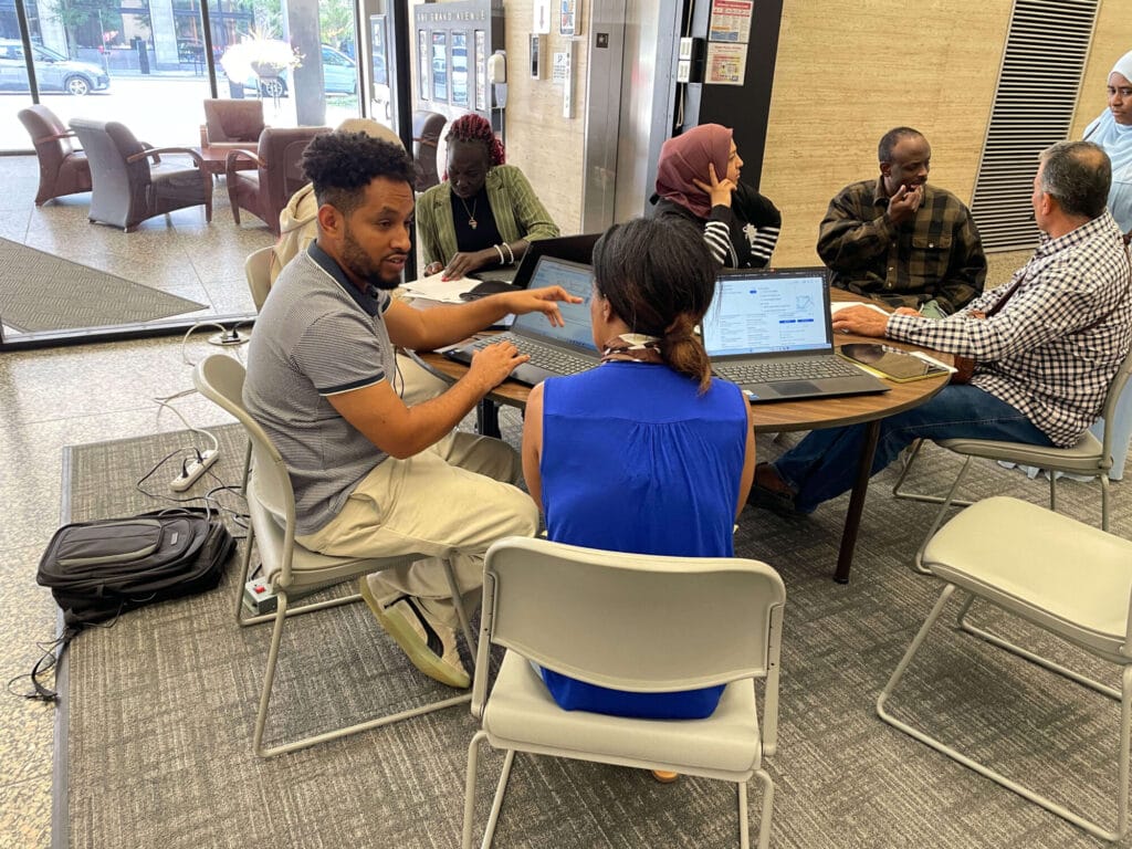 A group of people seated around a table, engaged in a discussion with laptops open in front of them. The setting appears to be a public or communal space, possibly a lobby or common area, with glass doors and windows in the background.